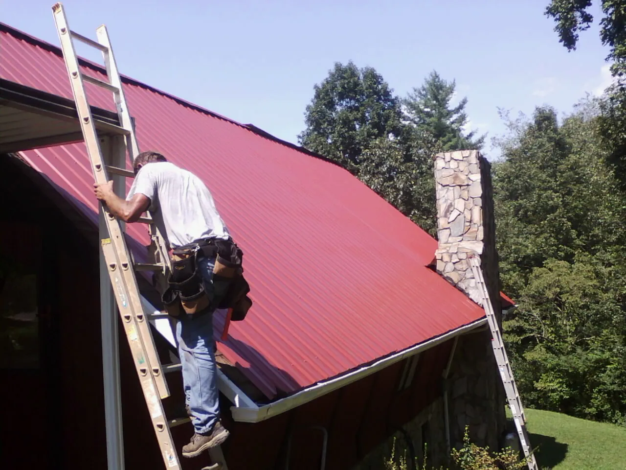 Roofer on a ladder beside a red metal roof and stone chimney — Soffit & Fascia Repair by Roofing Brothers in Prairie View